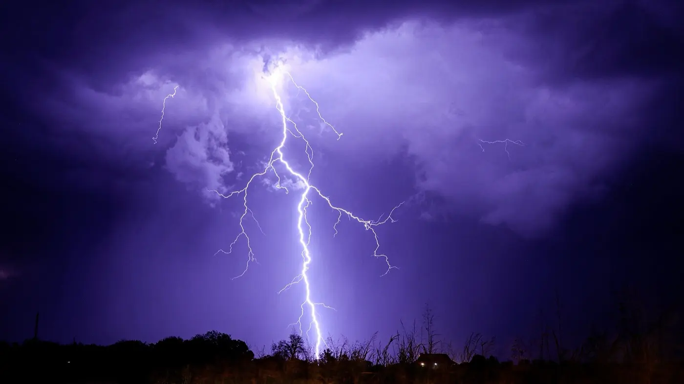 紫色に照らされた夜の雲から、白く輝く稲妻が地面へ落ちている写真