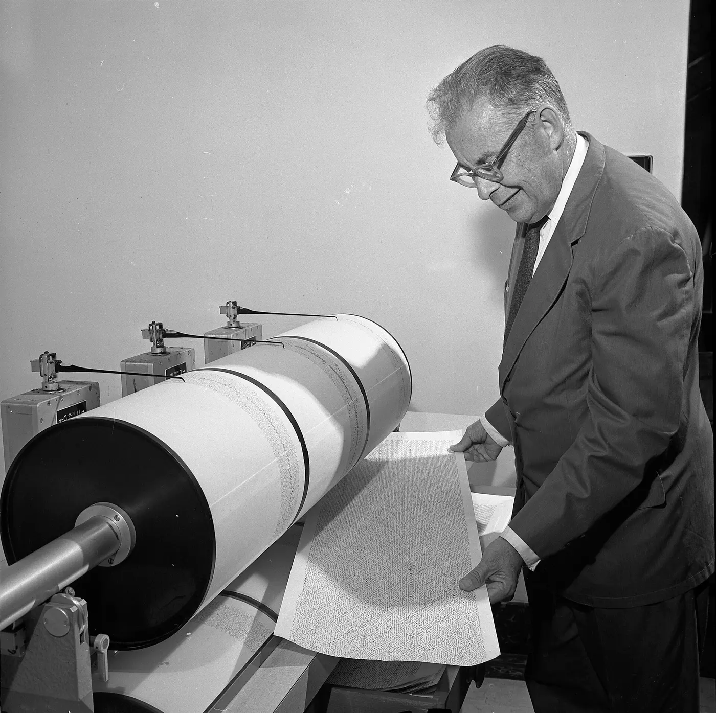 A man in glasses holding open a seismograph record beside a drum wrapped in recording paper