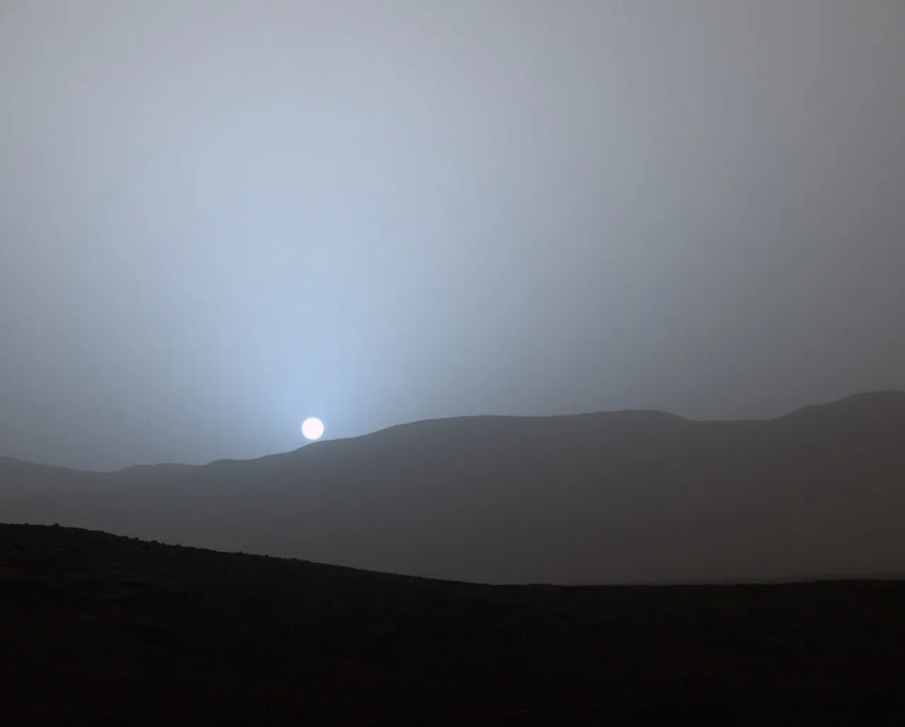 A small white Sun setting behind a distant ridge under a grayish sky, with a pale blue glow only around the Sun
