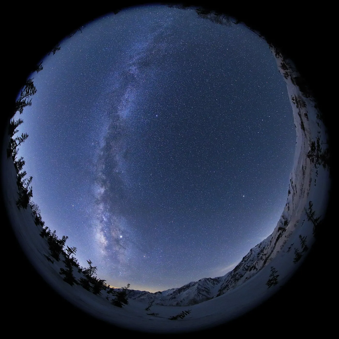 A fisheye photo of a circular starry sky, ringed by snowy mountains and trees, with the white Milky Way running vertically across it