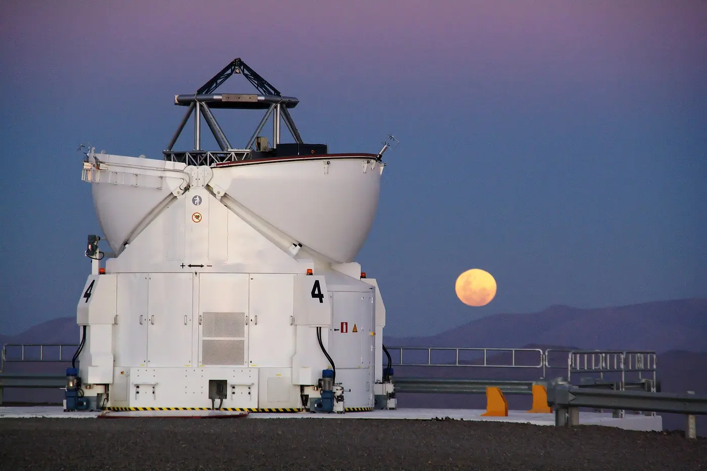 A reddish full Moon low on the horizon in a twilight sky, beyond a white telescope enclosure