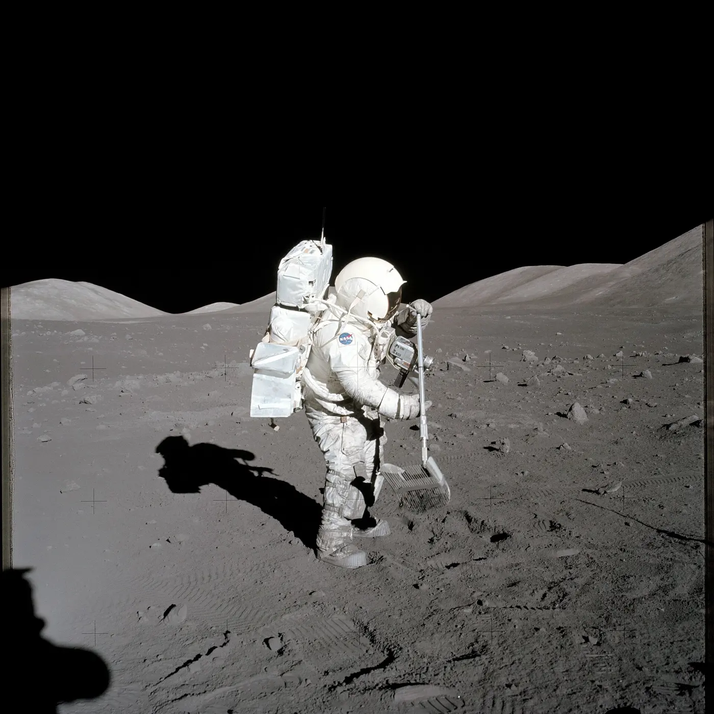 An astronaut in a spacesuit on the lunar surface, collecting rocks with a long-handled, rake-like tool