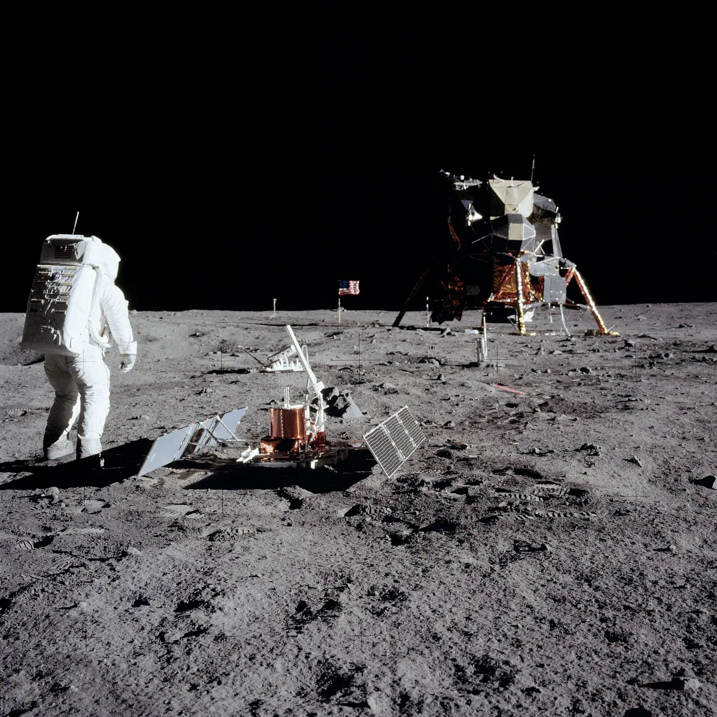 An astronaut in a spacesuit standing beside a seismometer package with solar panels spread out on both sides, on the lunar surface