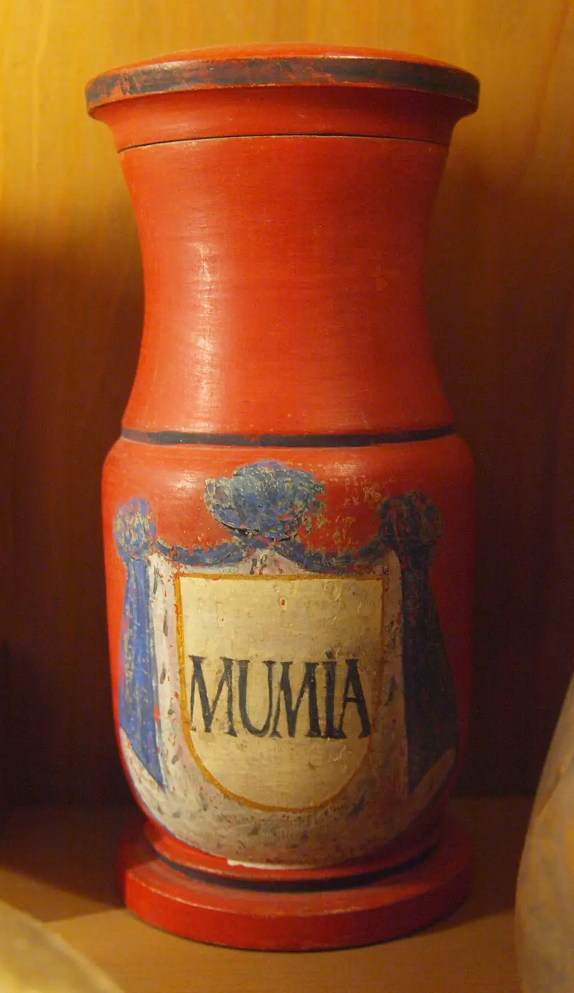 A red cylindrical container with “MUMIA” written inside a white shield-shaped panel