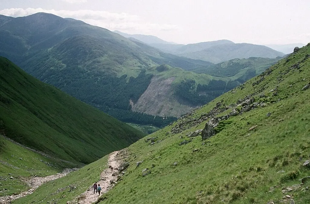 A narrow trail winding down a green mountainside, with two figures walking along it