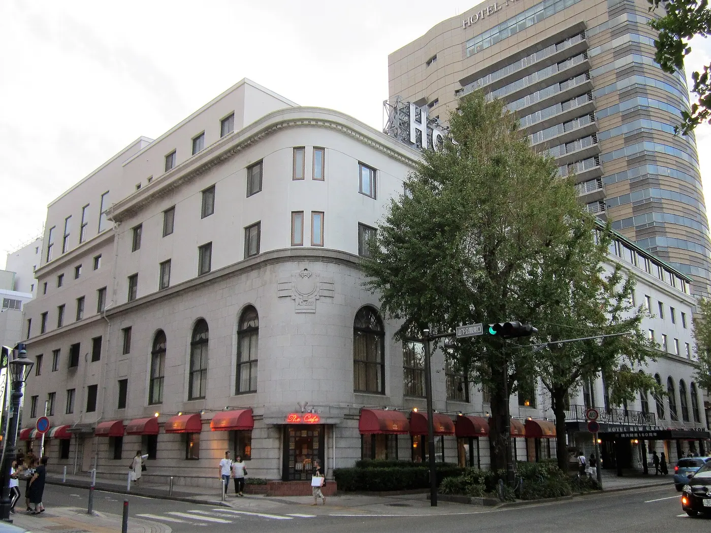 A cream-colored older building with arched windows and red awnings on a street corner, with a high-rise tower behind it