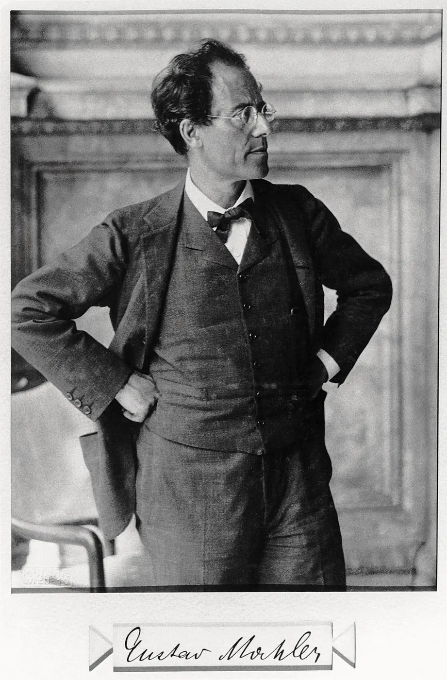 Black-and-white photo of a man in glasses standing with his hands on his hips, looking to the side, with his signature below