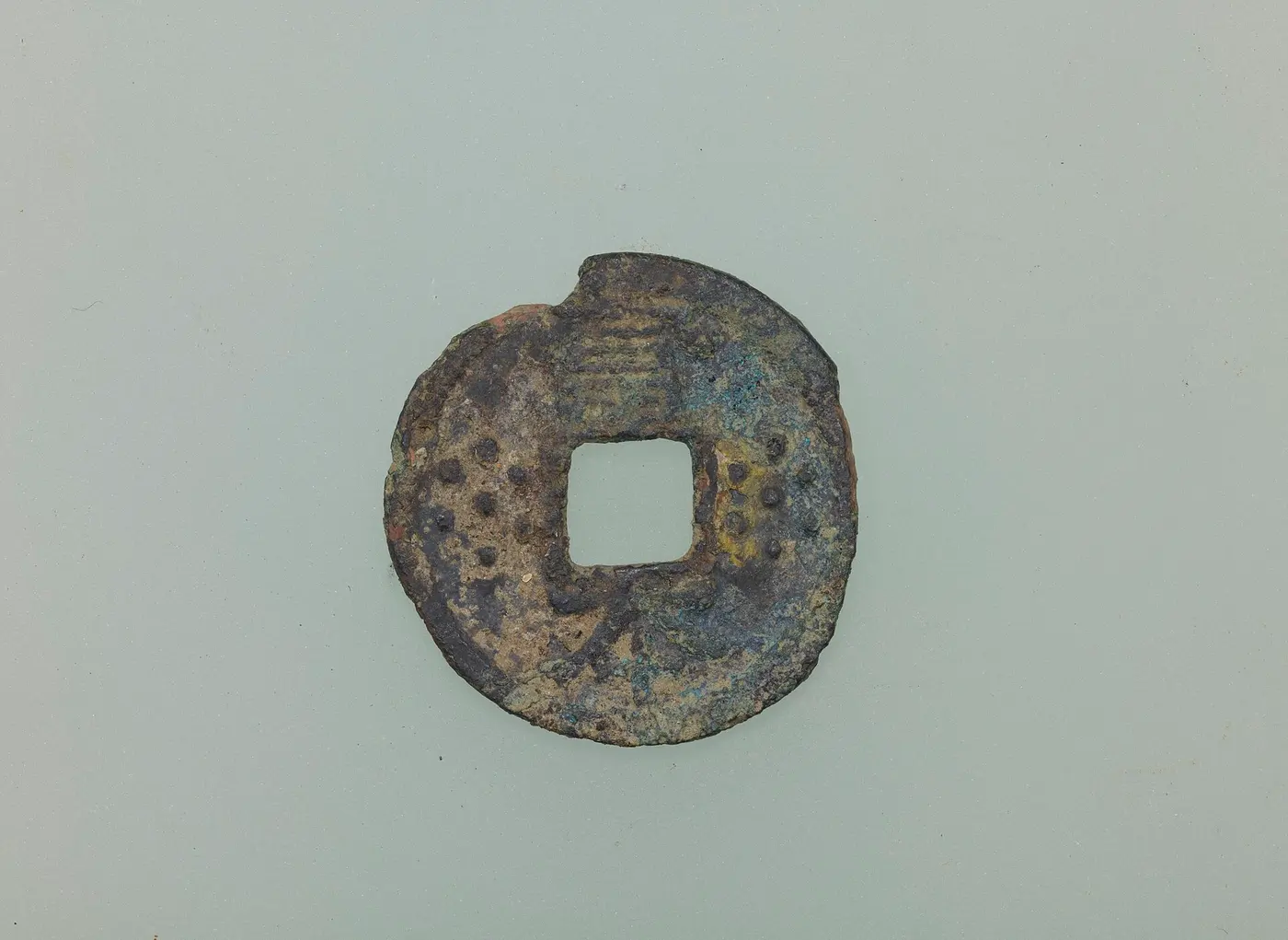 An old, corroded copper coin with a square hole in the middle and small dots lined up on either side of the hole