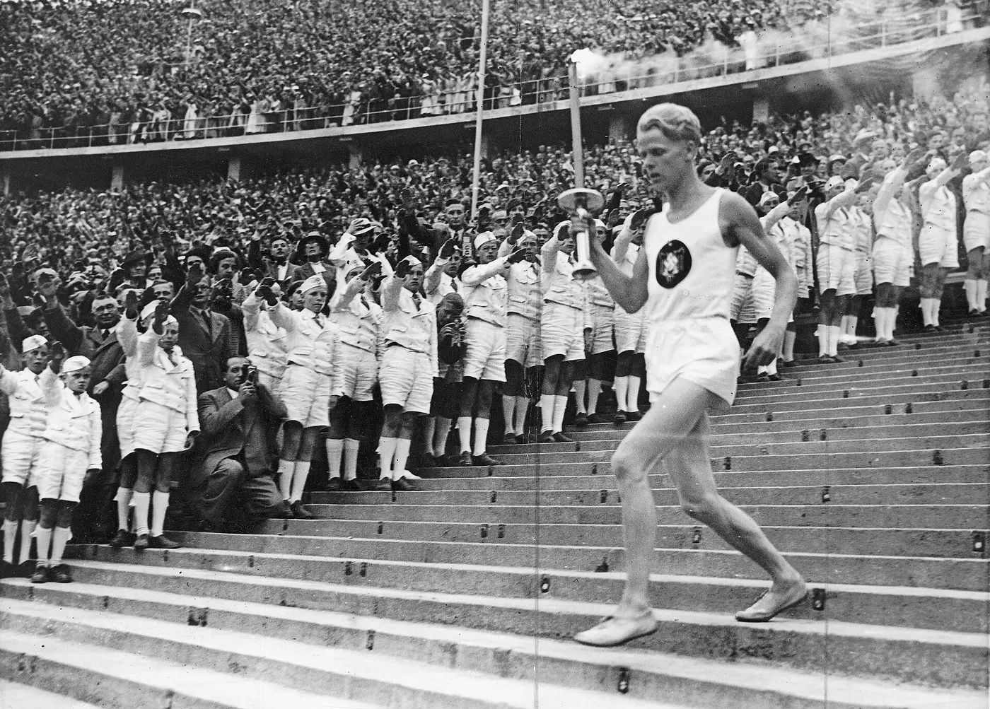 A runner in a white uniform holding a torch aloft as he runs down stadium steps before a huge crowd