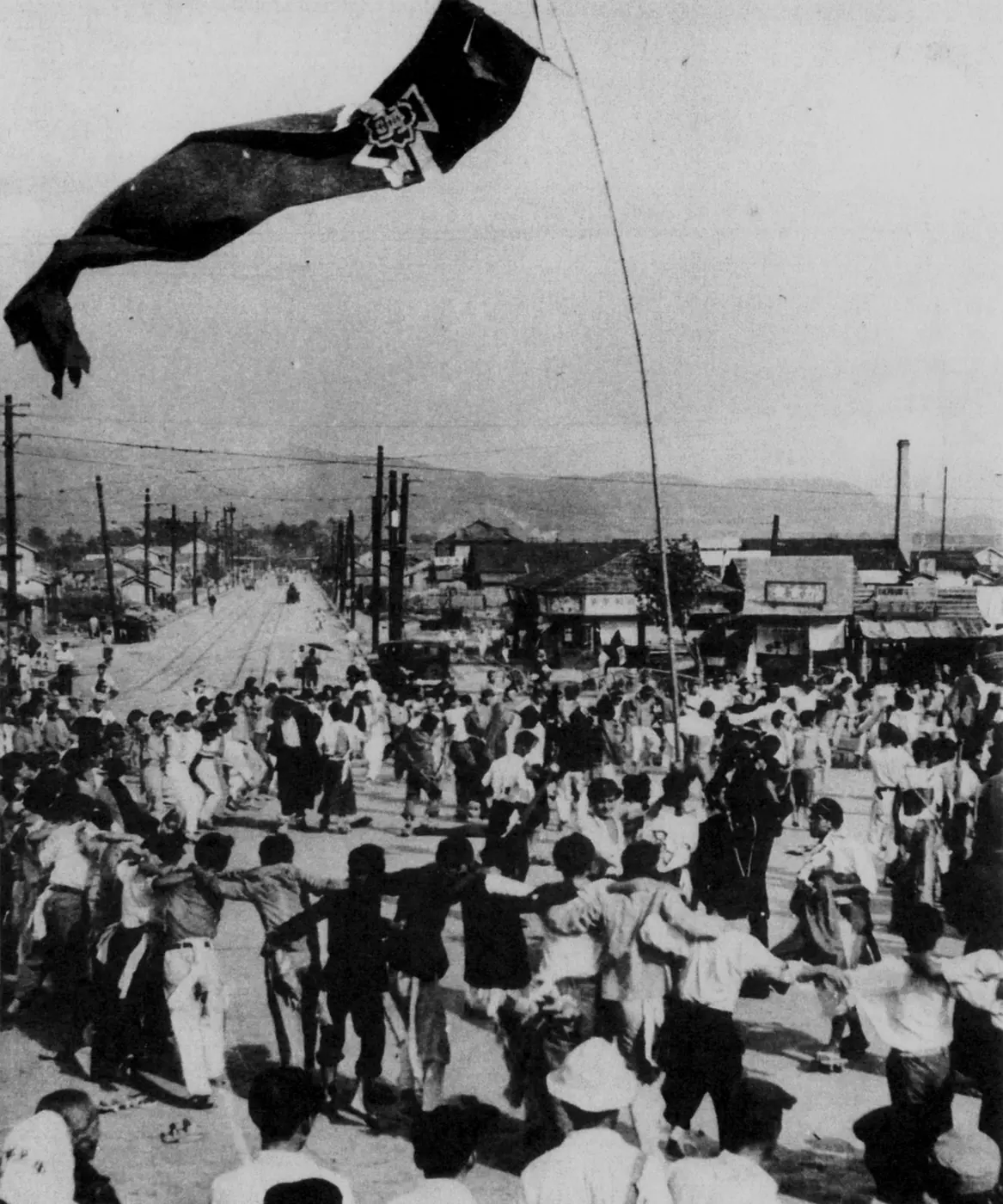 A black-and-white photograph of a crowd of students dancing in a ring with linked arms on a street, a large banner flying overhead