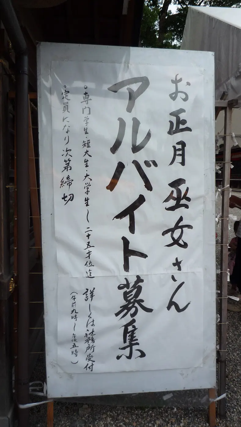 A brush-written sign standing in a shrine precinct, advertising part-time jobs as shrine maidens for the New Year