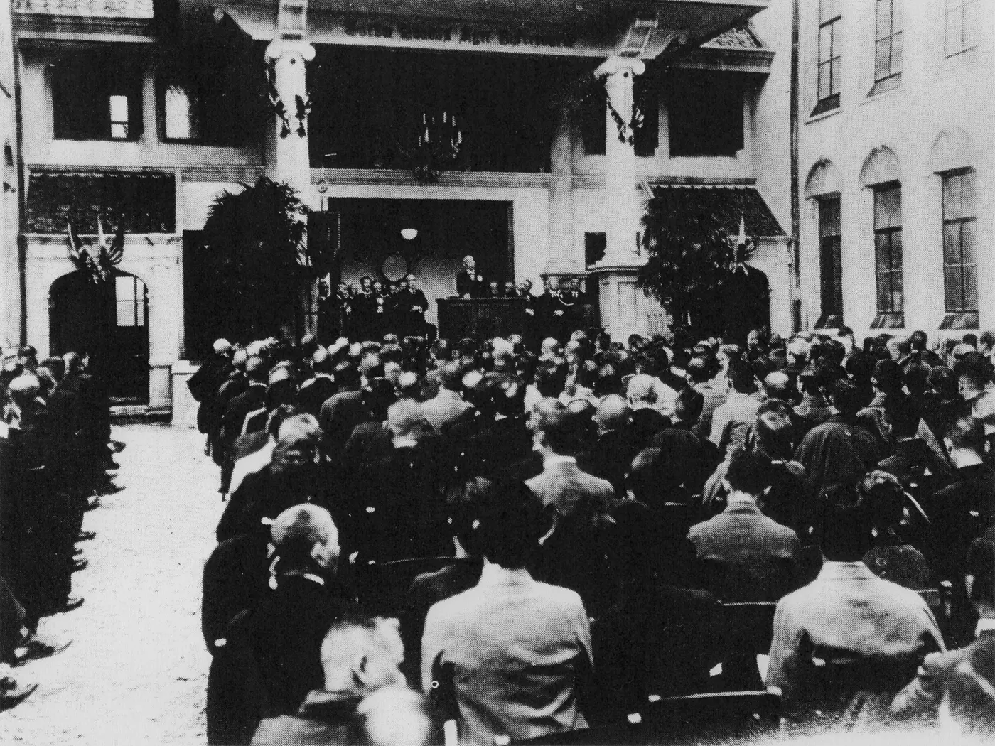 A black-and-white photograph of a large audience gathered in front of a Western-style building, with a man giving a speech at the front