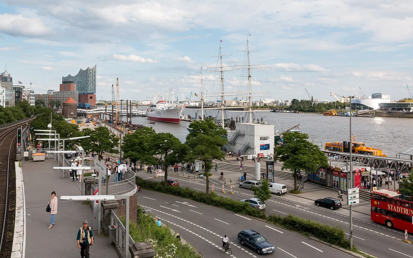A city view looking past a riverside road and trees to a harbor with a tall sailing ship and large vessels moored, and a glass-fronted building