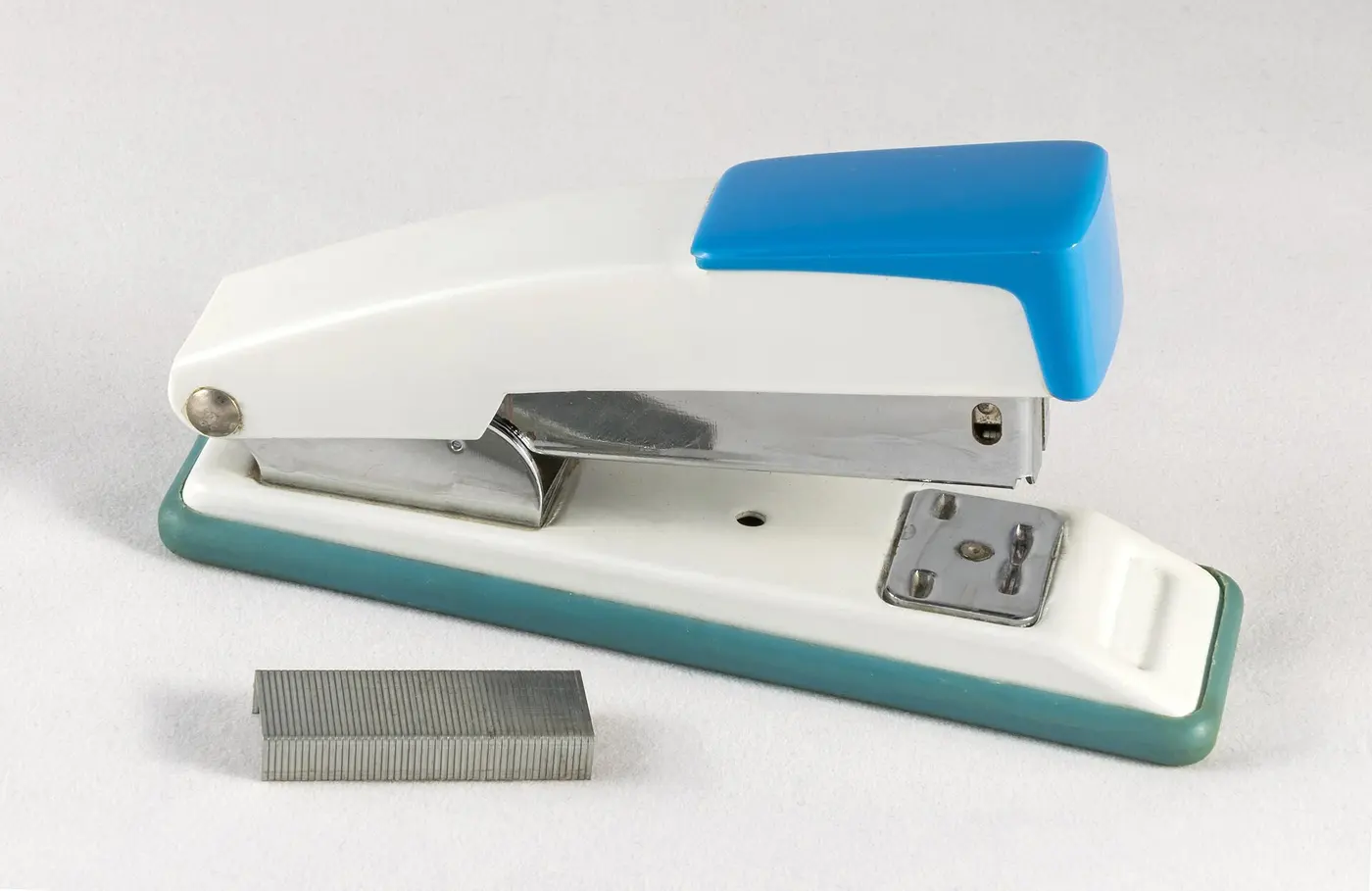 A white and blue plastic desk stapler next to a strip of staples