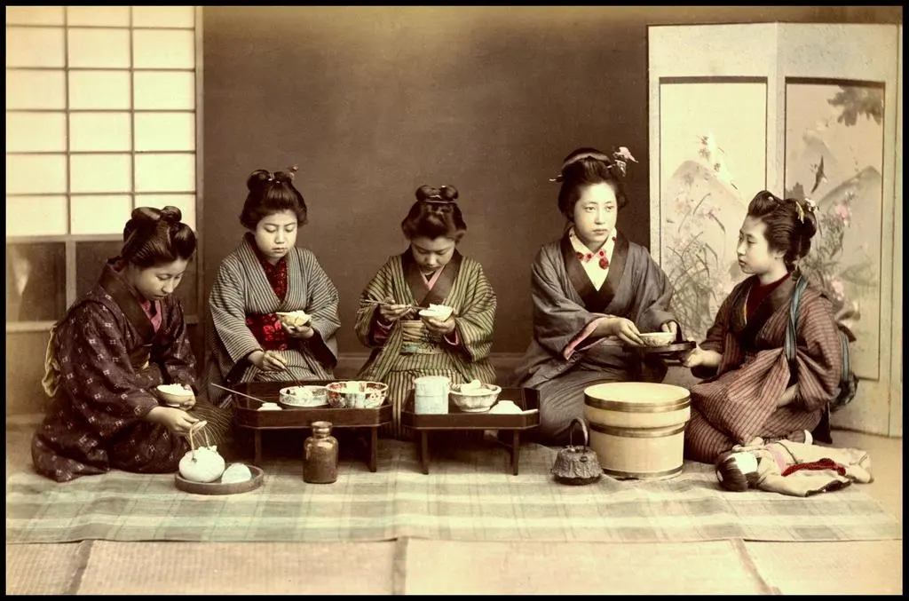 A hand-colored photograph of women in kimono sitting on tatami mats, each eating rice from a small tray table in front of her