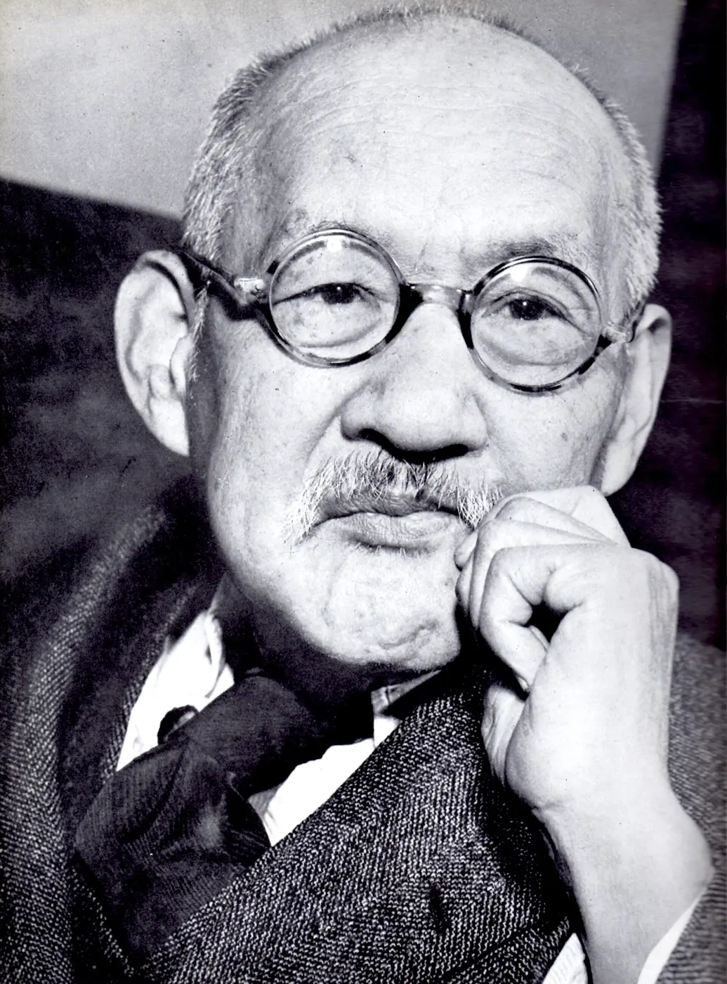 A black-and-white photo of an elderly man with round glasses and a mustache, resting his hand against his cheek