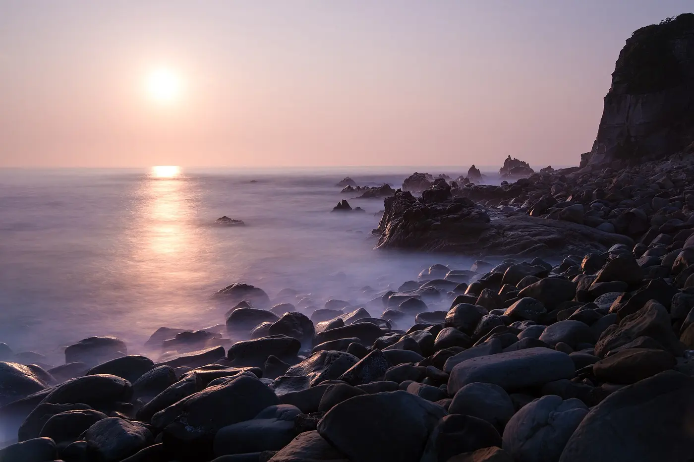 The sun rising in a pale purple sky beyond a rocky shore