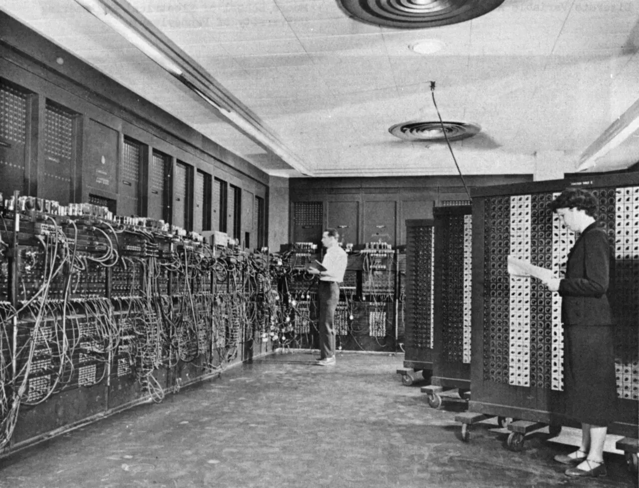 A black-and-white photo of a huge machine of wires and vacuum tubes lining the walls of a room, with two people working in front of it