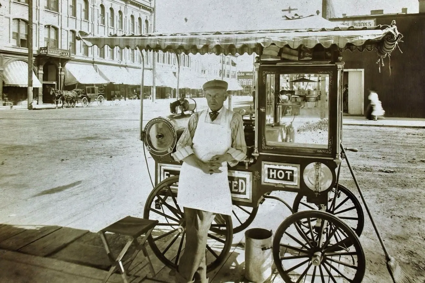 Black-and-white photo of a man in a white apron standing beside a wheeled cart carrying a glass-sided machine on a city street