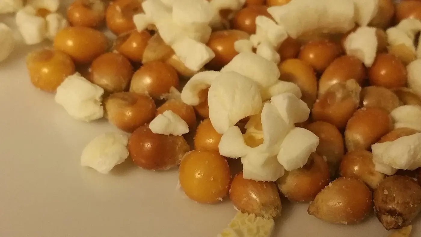 Glossy yellow popcorn kernels scattered on a white surface alongside white popped pieces