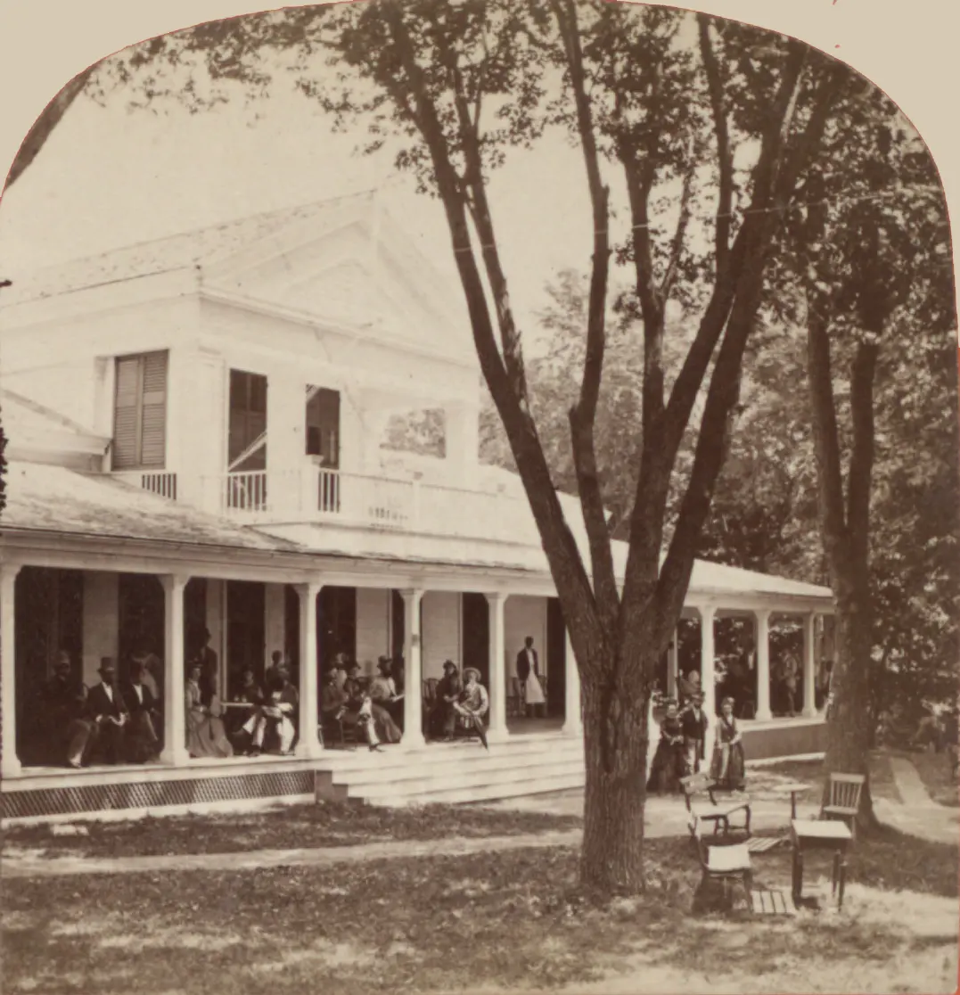 An old photo of a white wooden building among trees, with guests gathered on the veranda