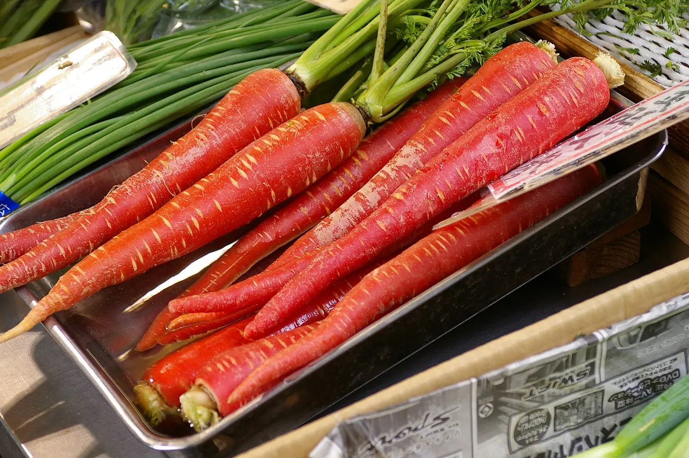 A metal tray holding many long, slender, bright red carrots with their leafy tops