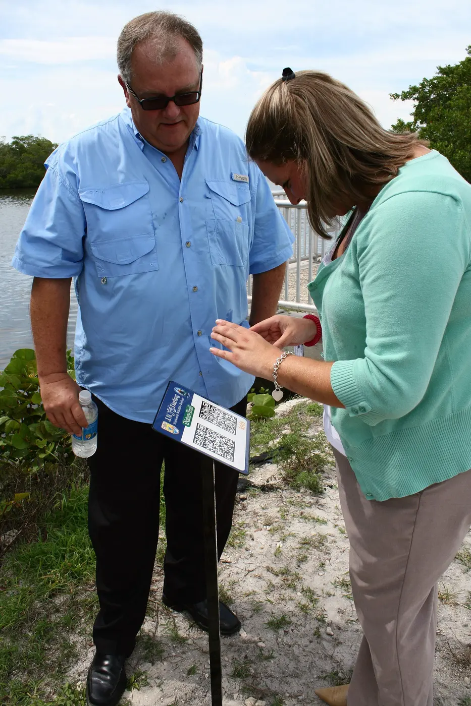 Two QR codes printed on a small outdoor information sign, with a person holding a smartphone nearby