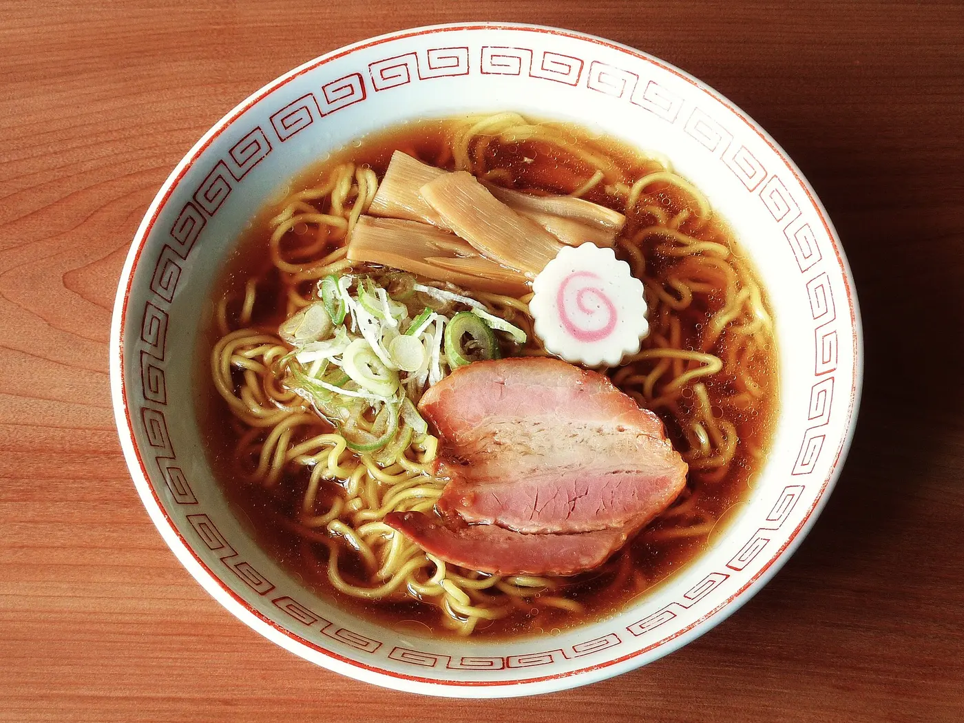 Ramen in a white bowl with a meander pattern, with soy-colored broth, wavy noodles, chāshū pork, bamboo shoots, narutomaki fish cake and green onion