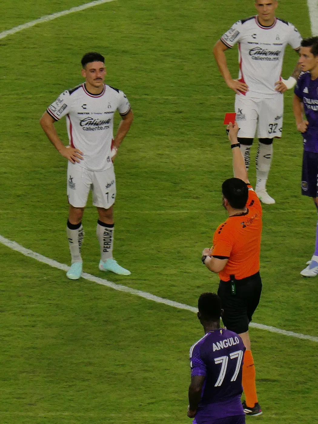 A referee in an orange shirt holds a red card high toward a player in a white uniform