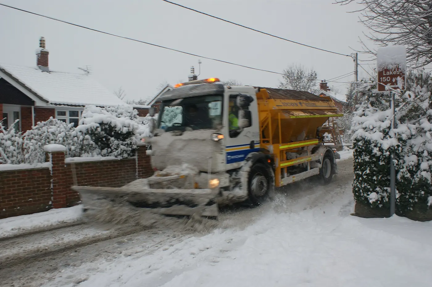 A truck with a snowplow blade at the front and an orange spreader on the back drives along a snow-covered residential street
