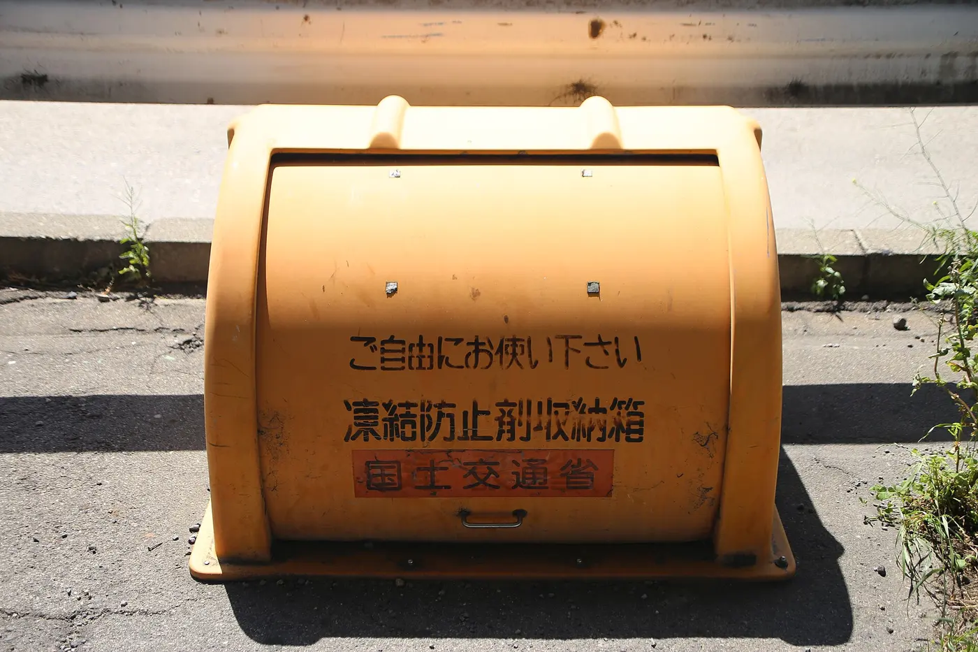 An orange, half-cylinder box beside a sidewalk, labeled in Japanese: Please use freely, de-icer storage box, Ministry of Land, Infrastructure, Transport and Tourism