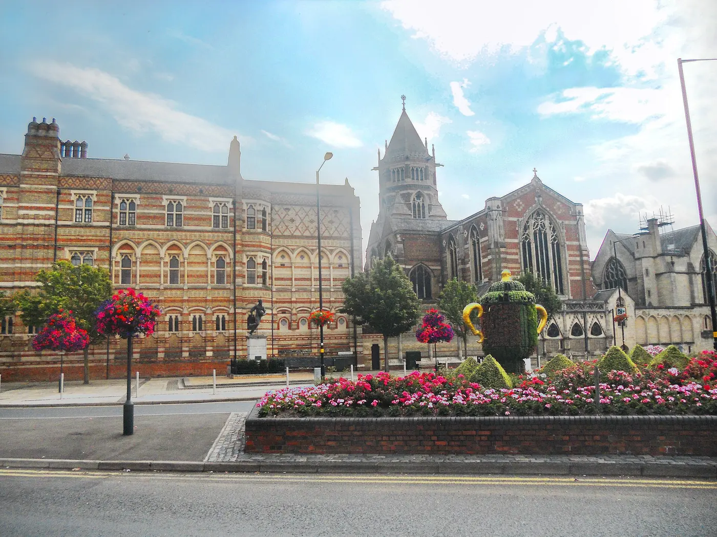 In front of a striped brick school building stands a bronze statue of a figure running with a ball, with a hedge clipped into the shape of a trophy in the flower bed in front
