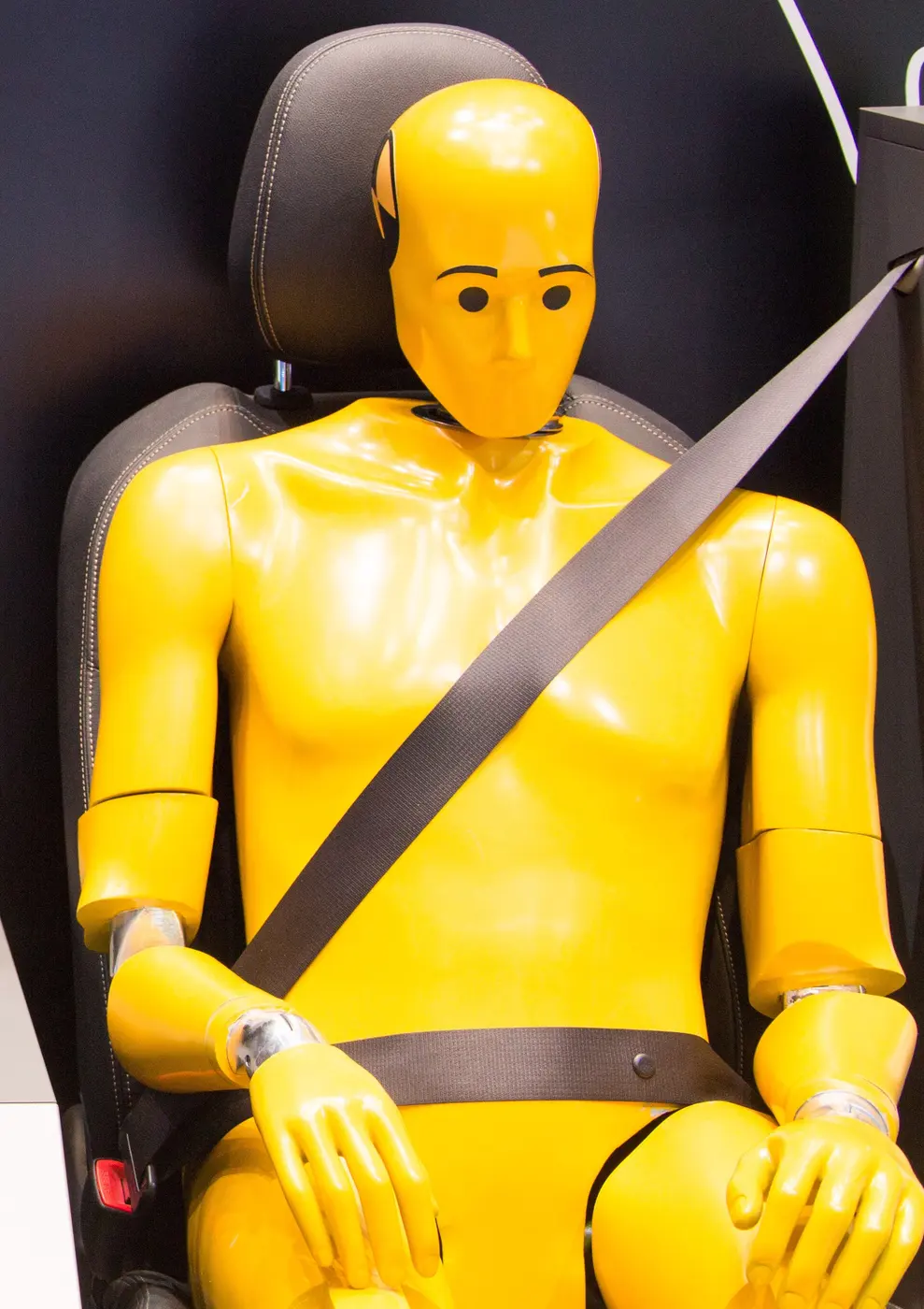 A yellow crash test dummy sitting in a seat, held by a diagonal belt over the shoulder and a belt across the lap