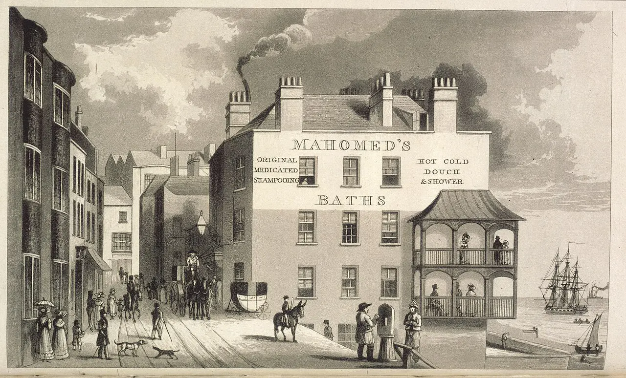 A print of a large building on a seaside street with “MAHOMED’S BATHS” on its wall, and carriages and people passing in front