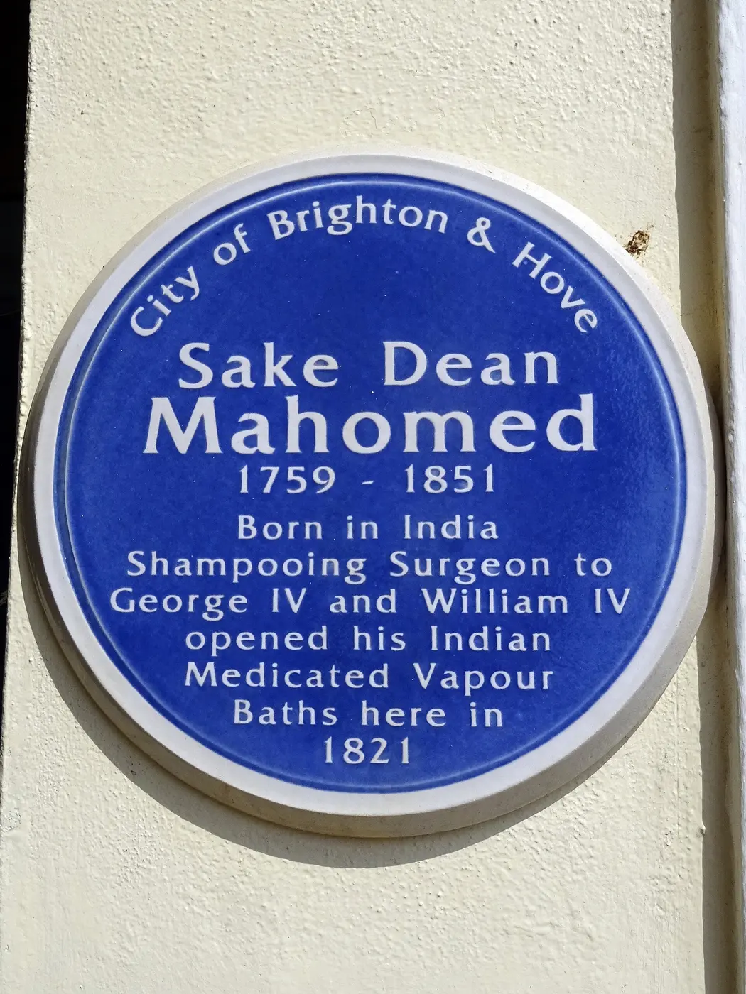 A round blue commemorative plaque on a white wall, bearing Sake Dean Mahomed’s name and noting in English that he opened baths there