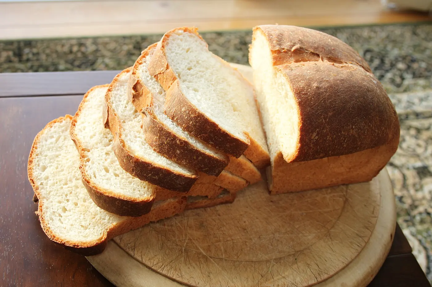 A white loaf with a rounded top, half cut into slices, on a wooden board