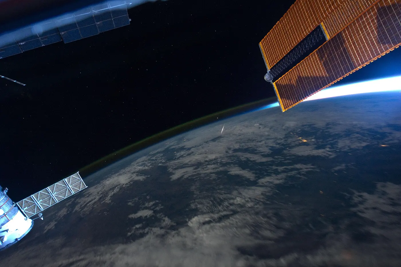 Earth at night seen between a space station’s solar panels, with a single meteor streaking just inside the edge of the atmosphere