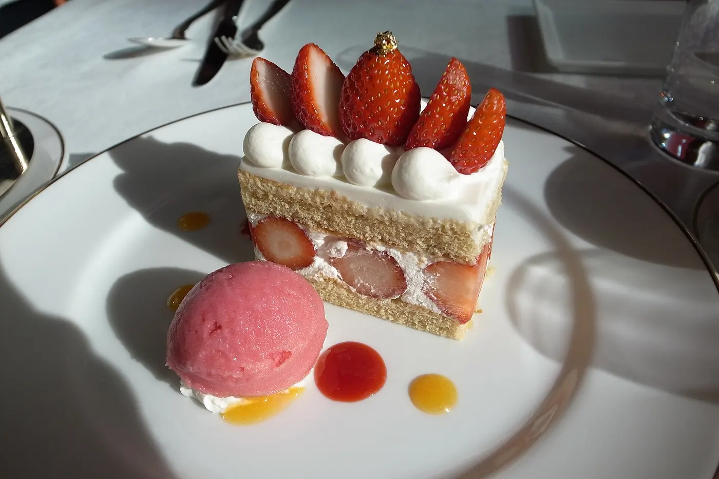 A rectangular slice of shortcake layered with sponge, cream and strawberries on a white plate, next to a scoop of strawberry sorbet