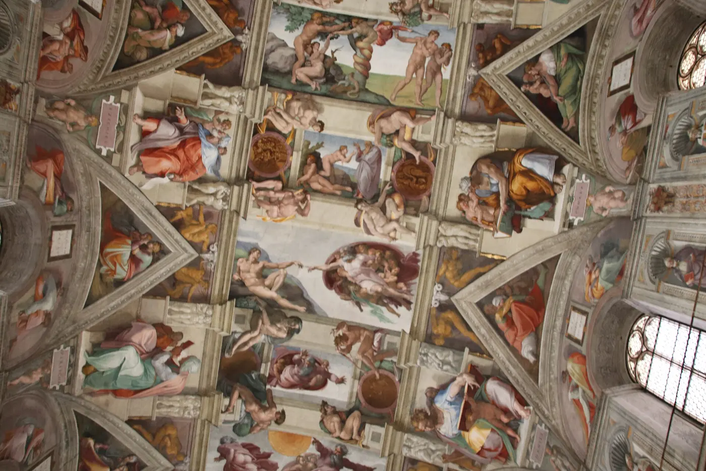 An arched ceiling covered in brightly colored figures and biblical scenes, with a reclining man and God reaching their fingers toward each other in the center
