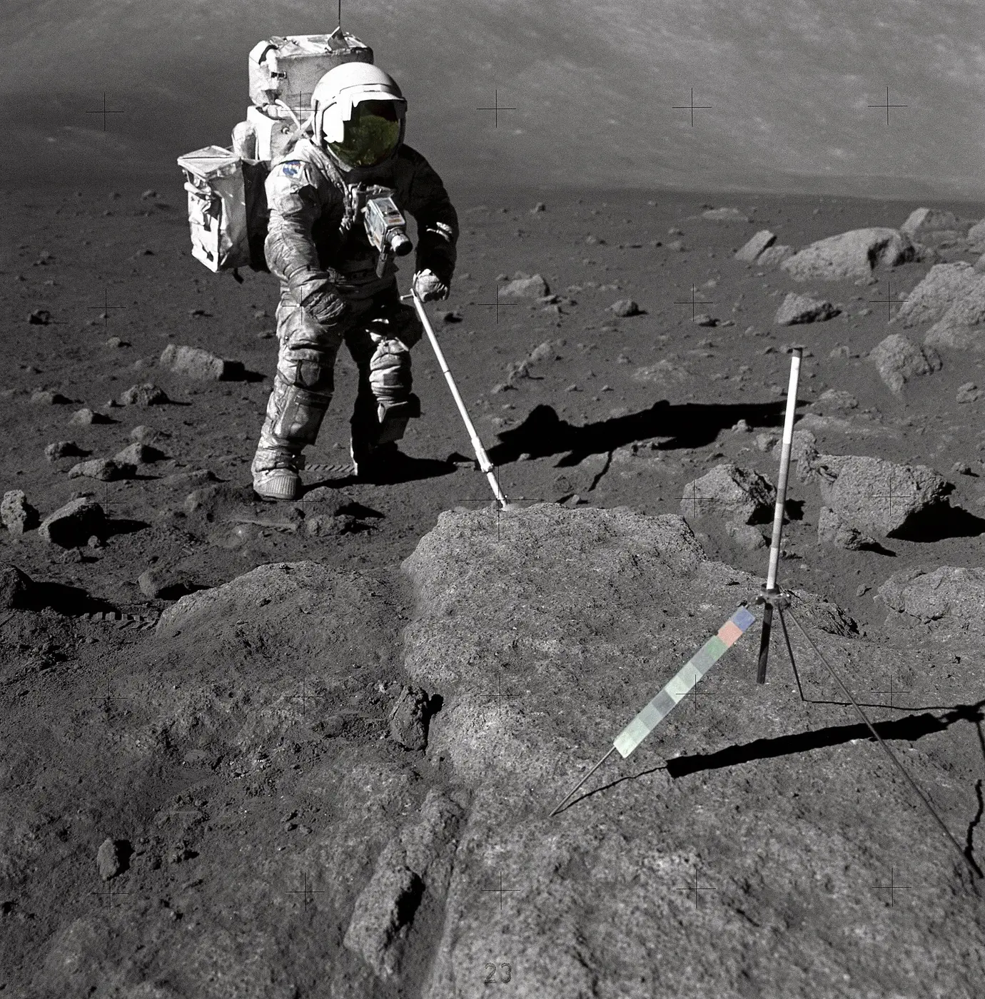 On the gray lunar surface, an astronaut with dust-smeared spacesuit legs and arms uses a long tool to collect rock samples