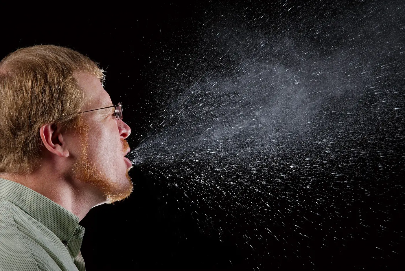 Side view of a man sneezing, with fine droplets spraying from his mouth in a cone
