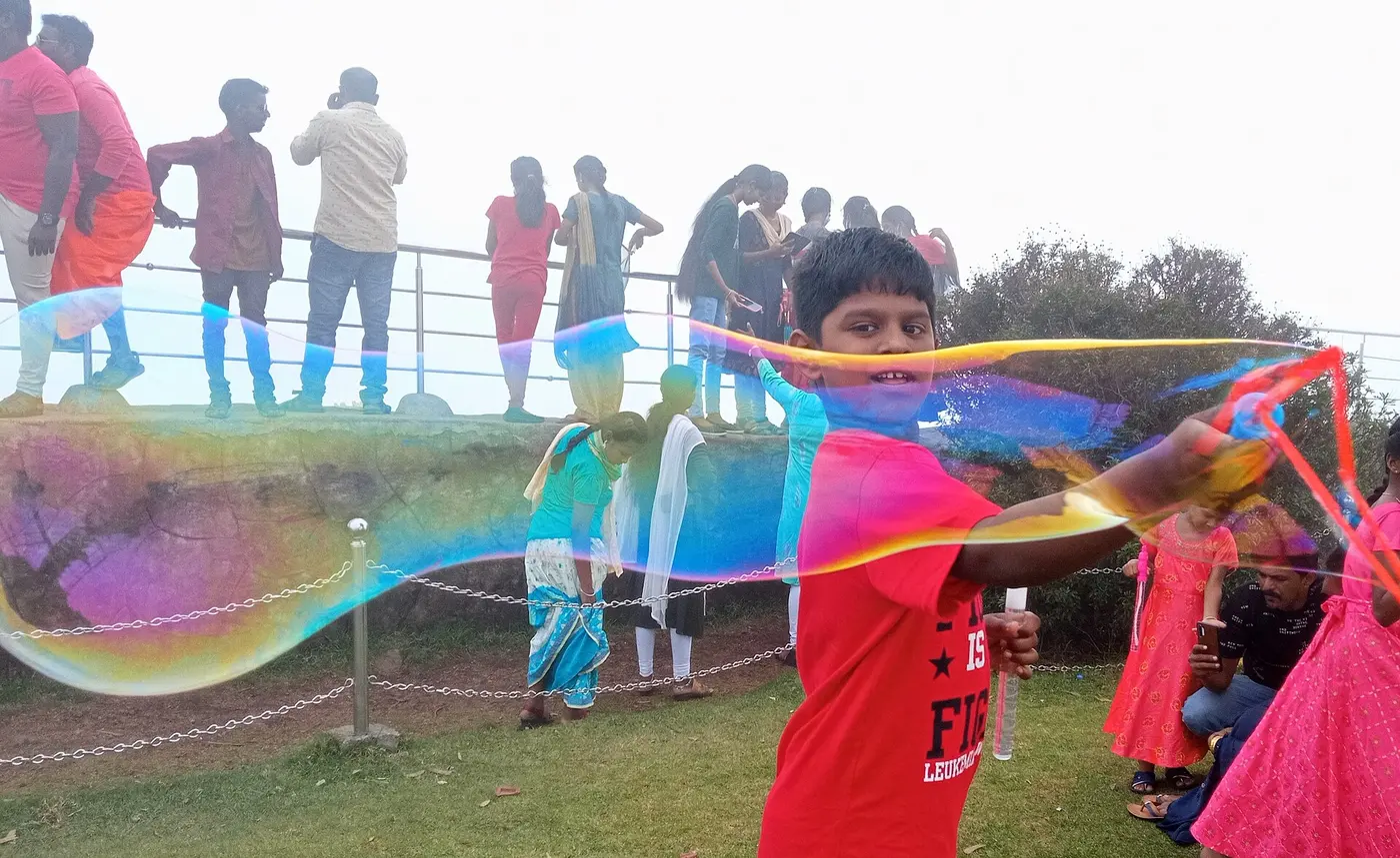 A boy outdoors making a large soap film, with bands of purple, blue and yellow appearing across the clear film
