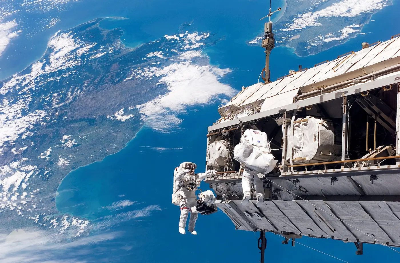 Two astronauts in spacesuits working outside the International Space Station, with Earth in the background