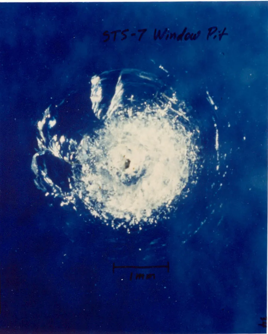 A round, white, shattered pit in the surface of bluish glass, next to a 1 mm scale