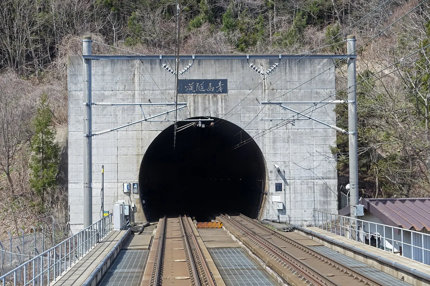 A tunnel portal bearing a sign reading “Seikan Tunnel,” with tracks leading into it that have three rails