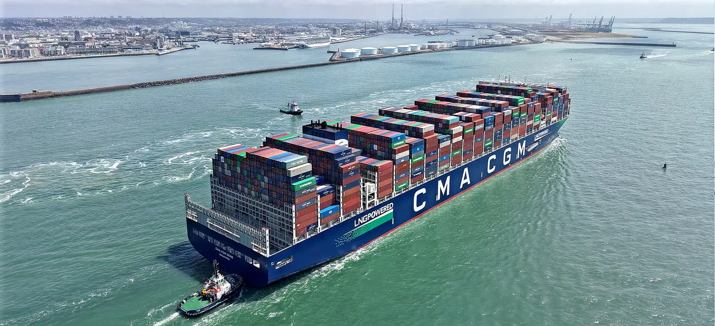A huge cargo ship stacked high with containers moves through a harbor entrance, escorted by a tugboat