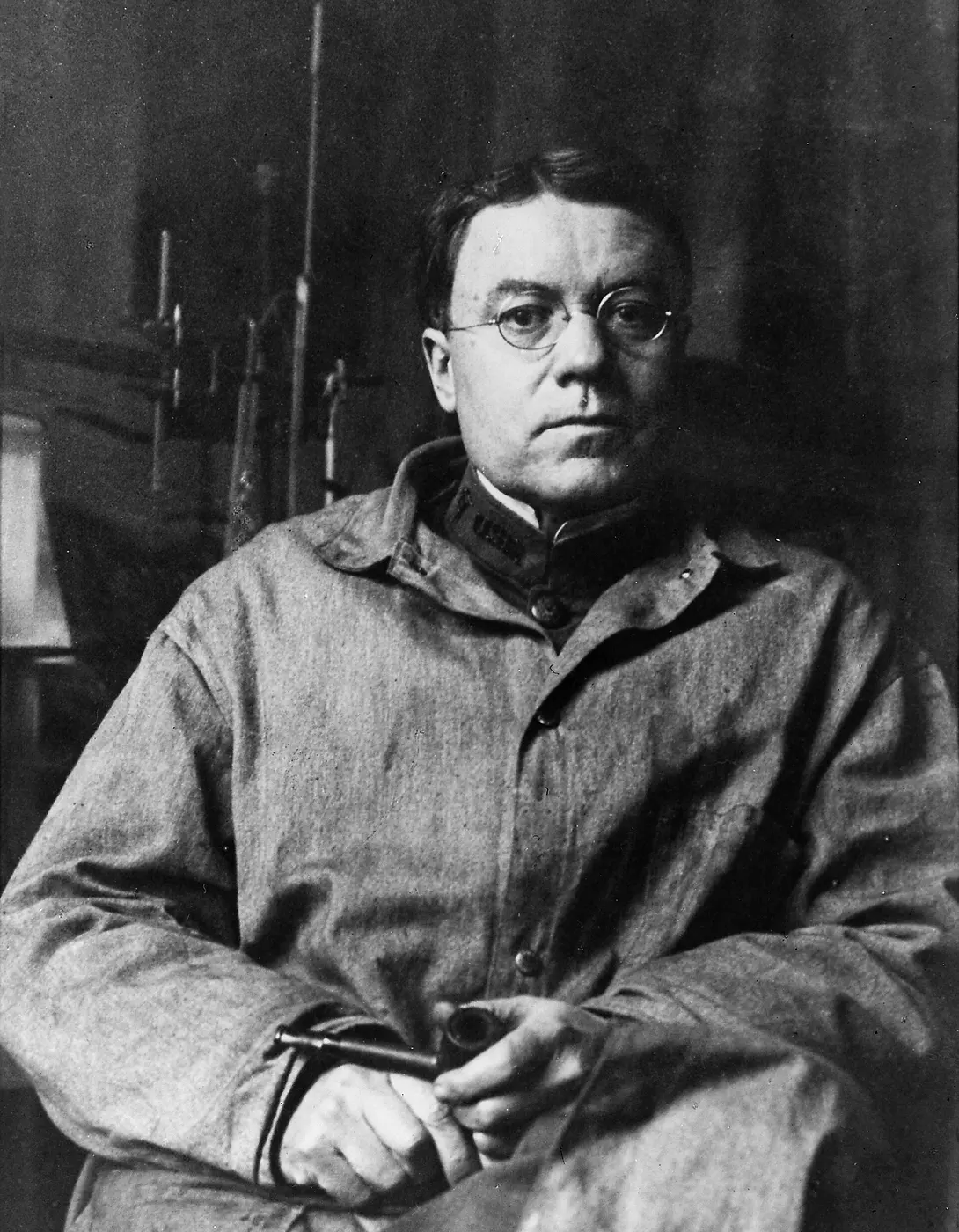 Black-and-white photo of a man in glasses and a work-coat-like jacket, seated in a chair