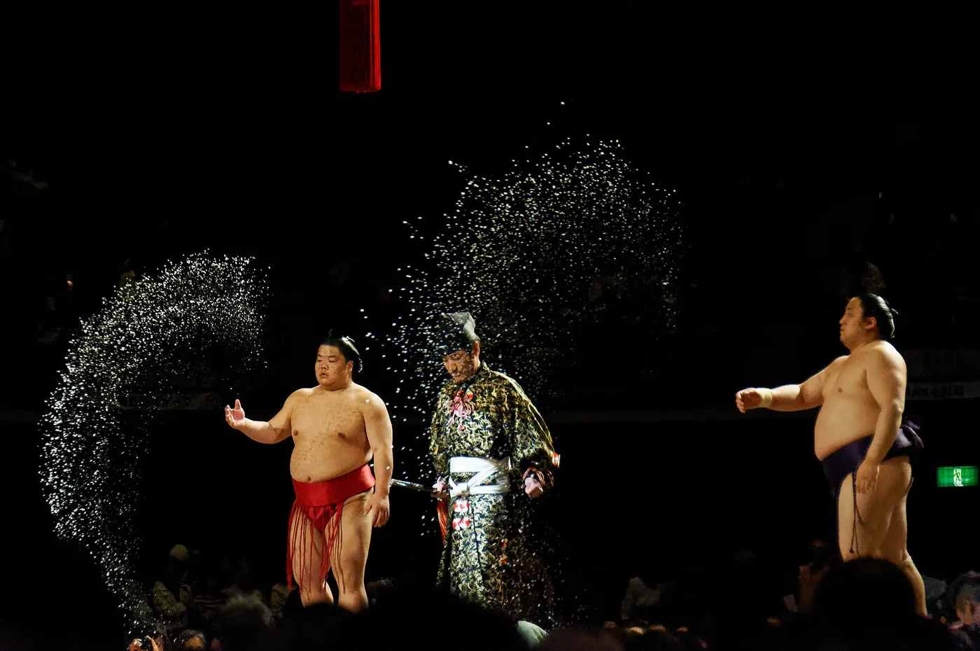 In a dark arena, two wrestlers on either side of a referee throw salt, the white grains arcing through the air