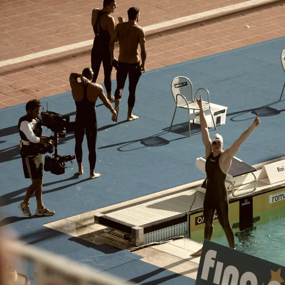 At the poolside, a swimmer in a black full-body suit raises both arms in triumph, with other swimmers in similar suits behind him