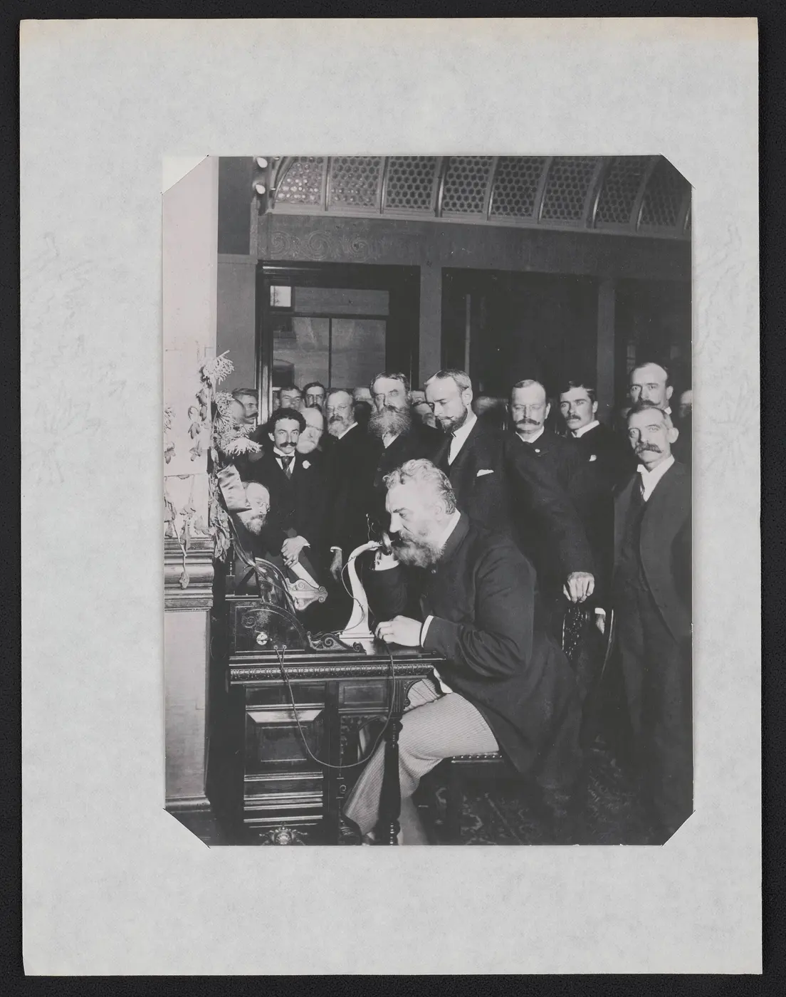 Black-and-white photo of a white-bearded man speaking into a mouthpiece on a desk while a crowd of men looks on